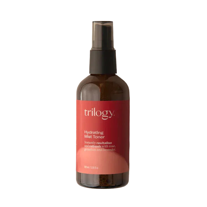 An amber glass bottle of Trilogy Hydrating Mist Toner with a black spray nozzle, set against a soft beige background with faint water droplets visible.