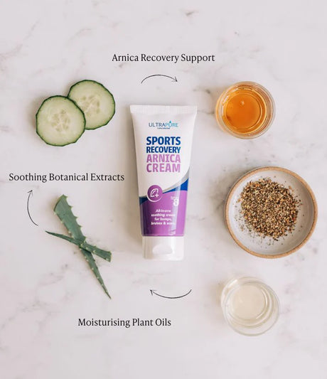 A flat-lay image featuring a tube of ULTRAPURE Laboratories Sports Recovery Arnica Cream on a marble surface, surrounded by botanical ingredients like cucumber slices, aloe vera, and plant oils. Text overlays highlight "Arnica Recovery Support," "Botanical Extracts," and "Moisturising Plant Oils."