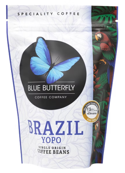 A stand-up pouch of Blue Butterfly Coffee Company Brazil Yopo single origin coffee beans. The packaging is primarily white with a large black circle containing a blue butterfly logo, blue text, and a colorful illustration of a parrot and tropical leaves on the right side.