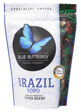 A stand-up pouch of Blue Butterfly Coffee Company Brazil Yopo single origin coffee beans. The packaging is primarily white with a large black circle containing a blue butterfly logo, blue text, and a colorful illustration of a parrot and tropical leaves on the right side.