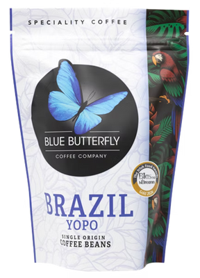 A stand-up pouch of Blue Butterfly Coffee Company Brazil Yopo single origin coffee beans. The packaging is primarily white with a large black circle containing a blue butterfly logo, blue text, and a colorful illustration of a parrot and tropical leaves on the right side.