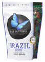 A stand-up pouch of Blue Butterfly Coffee Company Brazil Yopo single origin coffee beans. The packaging is primarily white with a large black circle containing a blue butterfly logo, blue text, and a colorful illustration of a parrot and tropical leaves on the right side.