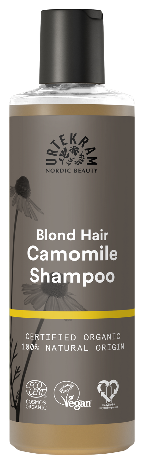 A tall bottle of Urtekram Camomile Shampoo for blond hair with a grey and brown label featuring camomile flower illustrations and a thin yellow horizontal stripe. The label indicates it is certified organic, 100% natural origin, and carries Vegan and Ecocert COSMOS Organic certifications.