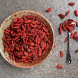 Dried goji berries in a rustic ceramic bowl with a silver spoon on a textured stone surface.