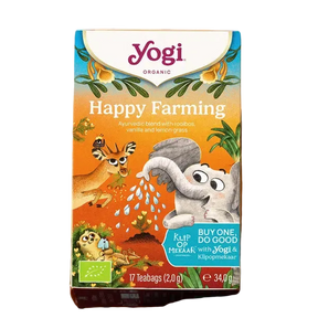 A box of Yogi Organic Happy Farming tea sits on a wicker tray beside a cup of tea. A plate with vanilla pods and a flower is nearby. The scene feels cozy and inviting.