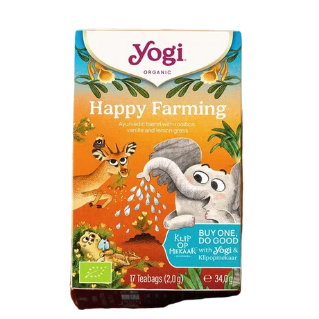 A box of Yogi Organic Happy Farming tea sits on a wicker tray beside a cup of tea. A plate with vanilla pods and a flower is nearby. The scene feels cozy and inviting.