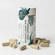 A cylindrical container of Moss Melts Organic Irish Sea Moss Capsules, a food supplement, surrounded by several scattered capsules on a white surface, featuring illustrative seaweed designs.