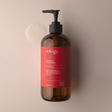 A large amber glass bottle of Trilogy Botanical Body Wash with a black pump dispenser, displayed against a soft beige background. 