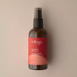 An amber glass bottle of Trilogy Hydrating Mist Toner with a black spray nozzle, set against a soft beige background with faint water droplets visible.