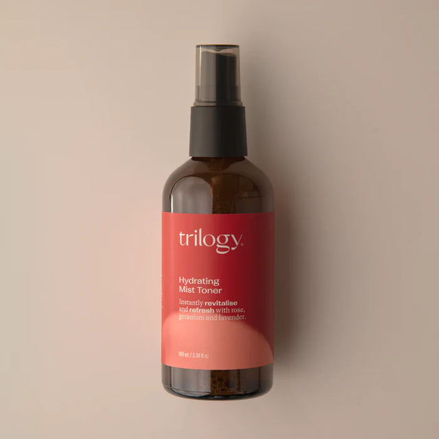 An amber glass bottle of Trilogy Hydrating Mist Toner with a black spray nozzle, set against a soft beige background with faint water droplets visible.