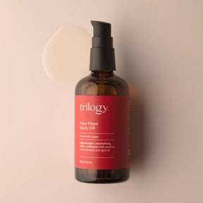 An amber glass bottle of Trilogy Pure Plant Body Oil with a black pump dispenser, set against a soft beige background.