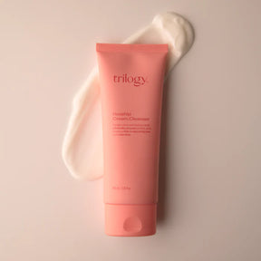 A tube of Trilogy Rosehip Cream Cleanser in a soft coral-pink matte finish, positioned over a thick, creamy white product swatch on a matching pastel background.