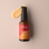 
A small amber glass bottle of Trilogy Vitamin C+ Super Serum with a black pump dispenser, set against a soft beige background with a creamy yellow product smear.