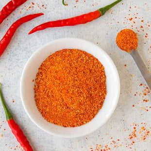 A white bowl filled with bright orange-red chili powder, surrounded by several whole red chili peppers and a small spoonful of the powder on a light gray textured surface.