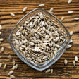 Overhead view of raw, shelled sunflower seeds in a square glass bowl set on a rustic wooden table.